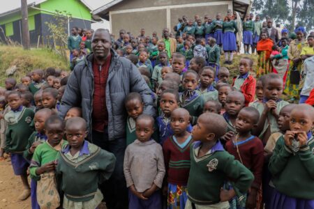 Un grand groupe d'enfants africains et un adulte se rassemblent devant une école rurale, illustrant l'esprit communautaire.