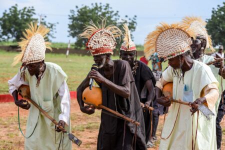 African musicians dressed in traditional attire perform with handmade instruments outdoors.