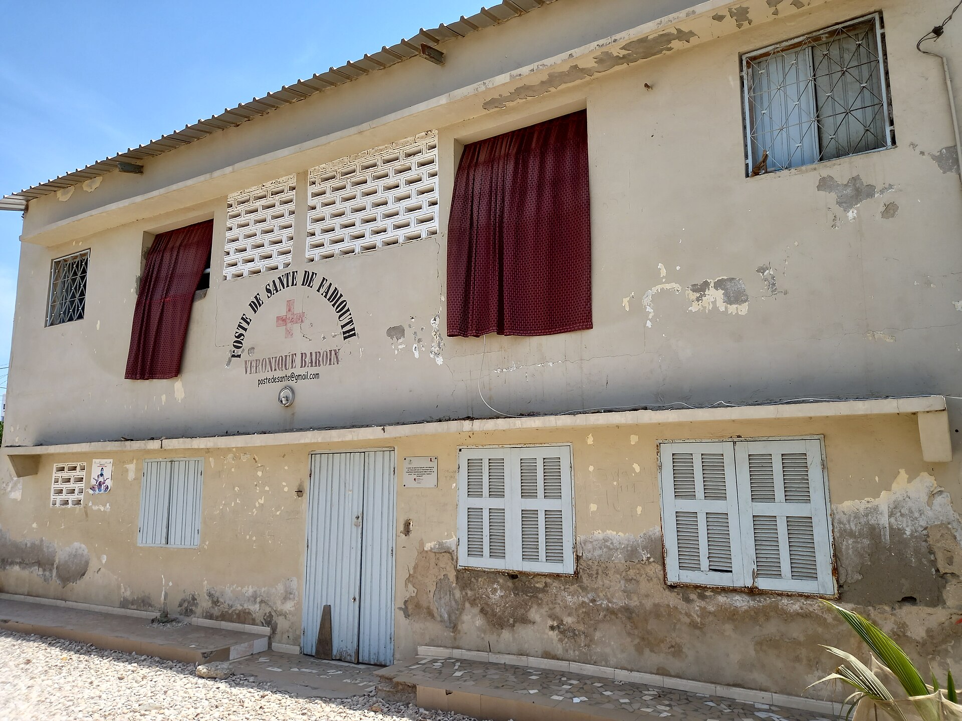 Façade défraîchie d’un poste de santé à étage, volets blancs et rideaux rouges aux fenêtres